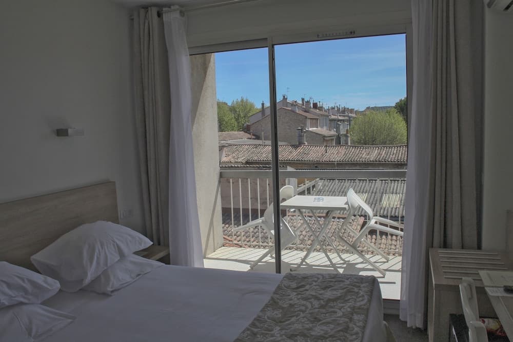 A cozy hotel room with a neatly made bed opens to a serene balcony overlooking tiled rooftops under a clear blue sky, ideal for travelers seeking relaxation and scenic views.