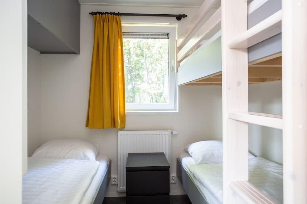 A cozy hostel room features bunk beds, a single bed, a vibrant yellow curtain framing a window view of greenery, and a minimalist design, ideal for budget travelers seeking comfort and simplicity.