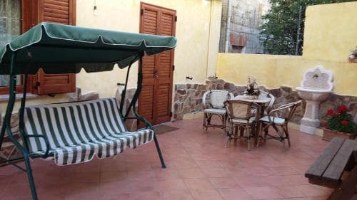A cozy courtyard patio features a striped swing under a green canopy, rustic wooden furniture around a table, and a stone fountain, evoking a serene and inviting atmosphere ideal for travelers seeking relaxation in a quaint outdoor setting.
