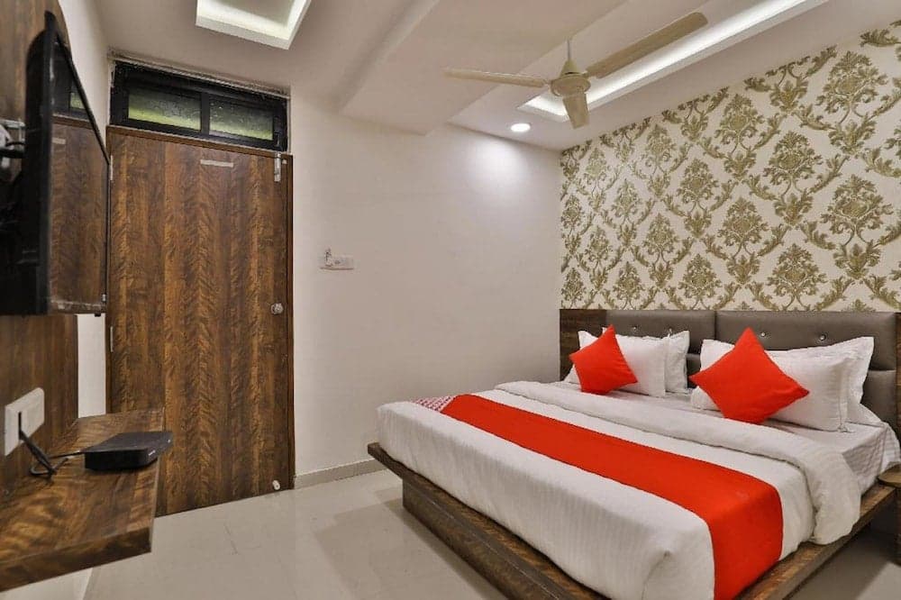 A cozy hotel room features a neatly made bed with red accents against patterned wallpaper, a wooden door, and a mounted TV, ideal for travelers seeking comfort and convenience.