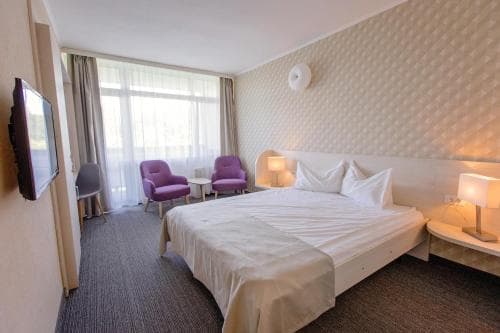 A cozy hotel room features a neatly made bed, purple armchairs by a large window, and modern amenities, creating a welcoming atmosphere ideal for travelers seeking comfort and relaxation.