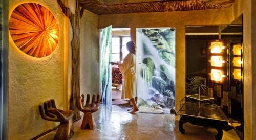 A serene spa interior features a person in a robe gazing at a waterfall mural, surrounded by rustic wooden furniture and warm lighting, ideal for relaxation and wellness retreats.