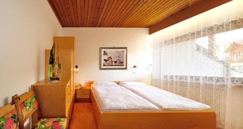 A cozy bedroom with wooden furnishings, lace curtains, and a framed picture creates a warm and inviting atmosphere, ideal for travelers seeking comfort in a quaint lodge or guesthouse.