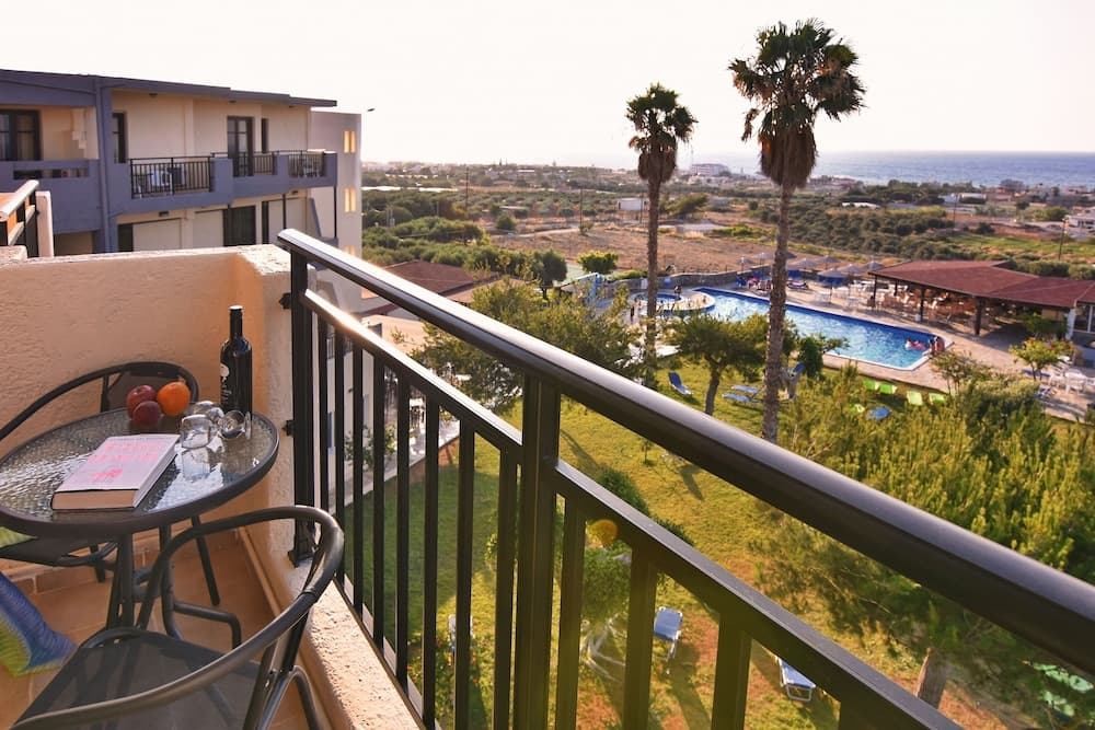 A serene hotel balcony overlooks a lush garden, palm trees, and a sparkling pool near the sea, ideal for travelers seeking relaxation and coastal views.