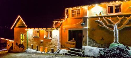 A cozy, warmly lit building adorned with festive lights and a mural of a tree stands invitingly in a snowy night scene, perfect for travelers seeking a charming winter getaway.