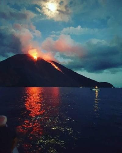A dramatic volcanic eruption lights up the night sky over a serene body of water, creating a mesmerizing reflection on the surface, ideal for adventurous travelers seeking unique natural phenomena photography opportunities.