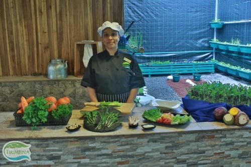 A chef stands proudly behind a vibrant display of fresh produce in an indoor garden setting, evoking a sustainable culinary experience ideal for eco-conscious travelers exploring innovative food practices.