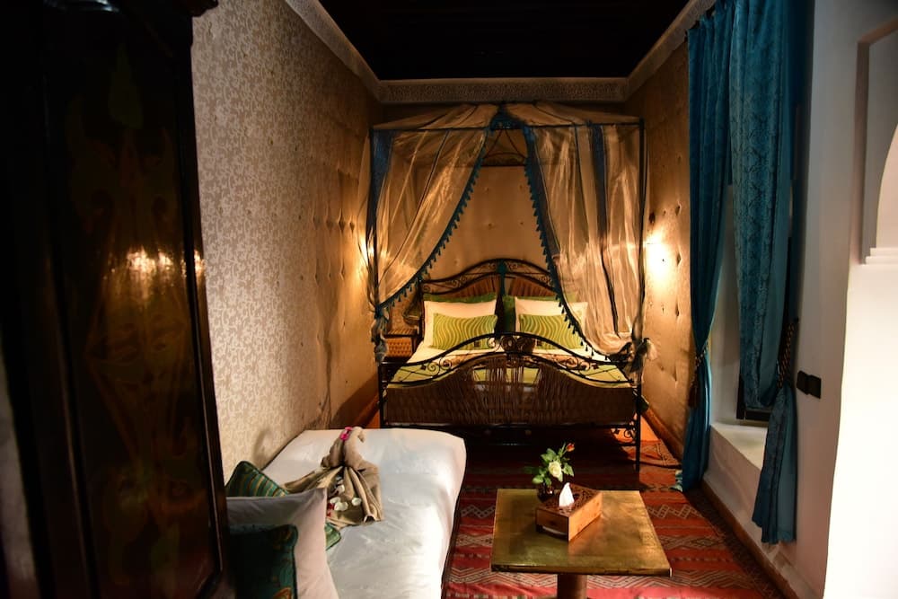 A cozy, warmly lit boutique hotel room features a canopy bed adorned with green pillows, patterned wallpaper, and a small table with flowers, ideal for travelers seeking a romantic and intimate getaway.