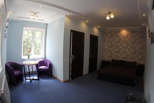 A cozy hotel room features a neatly made bed, purple armchairs by a sunlit window, and patterned wallpaper, creating a welcoming atmosphere ideal for travelers seeking comfort and relaxation.