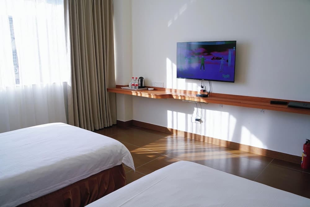 A cozy hotel room with two neatly made beds, a wooden desk featuring a mounted TV and refreshments, soft natural light filtering through sheer curtains, and a serene ambiance ideal for travelers seeking relaxation.