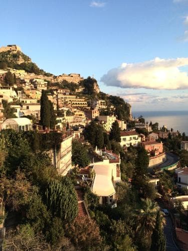 A picturesque coastal hillside town bathed in warm sunlight, featuring terraced buildings, lush greenery, and a distant castle, evokes a serene Mediterranean escape ideal for travelers seeking relaxation and scenic beauty.