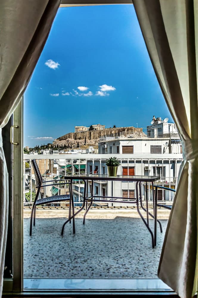 A serene balcony view framed by curtains showcases a vibrant blue sky and the iconic Acropolis, ideal for travelers seeking a picturesque urban retreat.