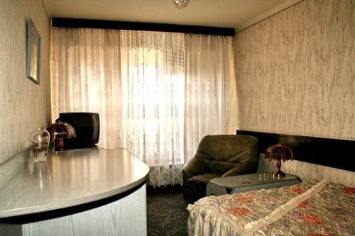 A cozy hotel room features a neatly made bed, a comfortable armchair by sheer curtains, a small desk with a vase, and a vintage TV, creating a warm and inviting atmosphere ideal for travelers seeking a quiet retreat.