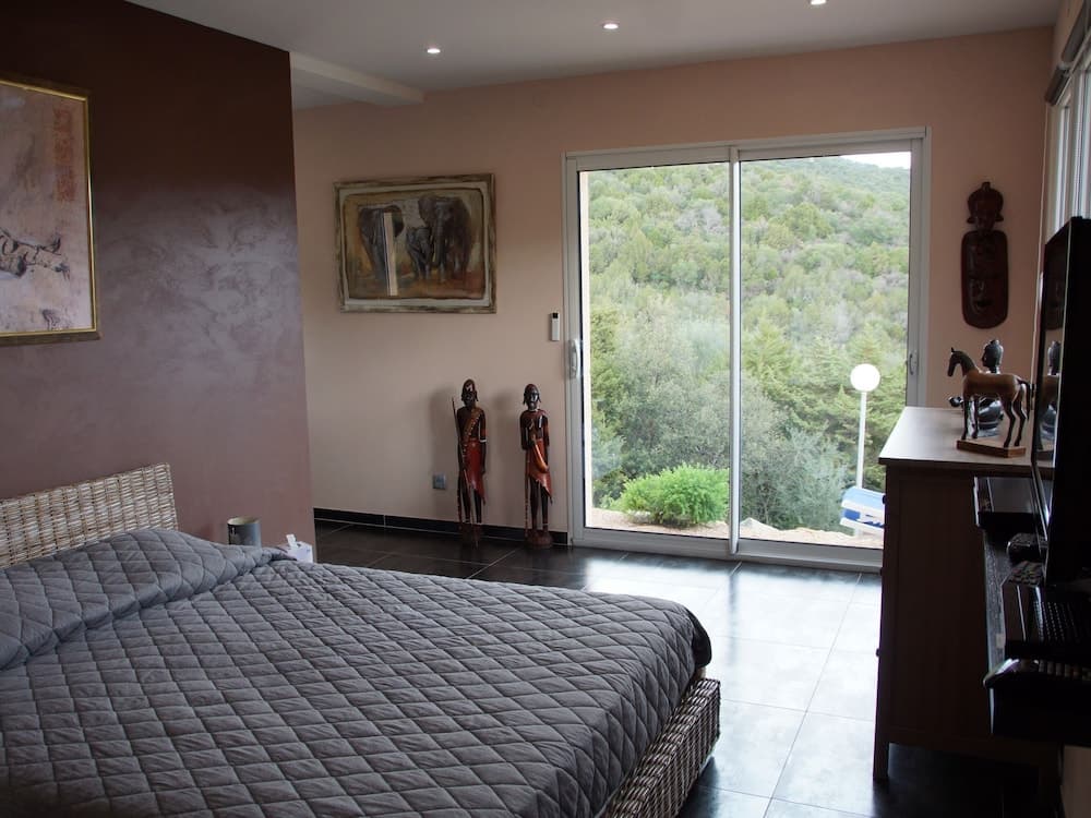 A serene bedroom with a cozy bed, cultural sculptures, and a scenic forest view through sliding glass doors, ideal for travelers seeking relaxation and nature-inspired tranquility.