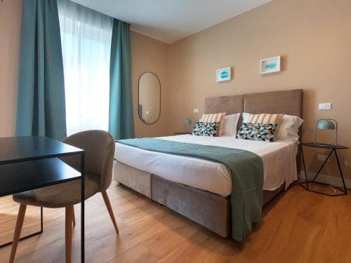 A cozy hotel room with a neatly made bed, wooden flooring, teal curtains, and modern furnishings creates a welcoming atmosphere ideal for travelers seeking comfort and relaxation.