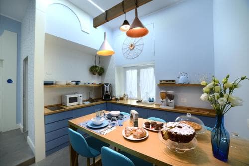 A cozy kitchen-dining area with blue cabinetry, warm pendant lights, and a wooden table set with pastries and flowers, ideal for travelers seeking a charming and inviting self-catering accommodation.