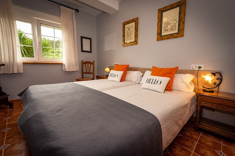 A cozy bedroom in Asturias, Spain, features a neatly made bed with orange and white pillows, wooden furniture, and a window view of lush greenery, ideal for travelers seeking a serene and inviting accommodation.
