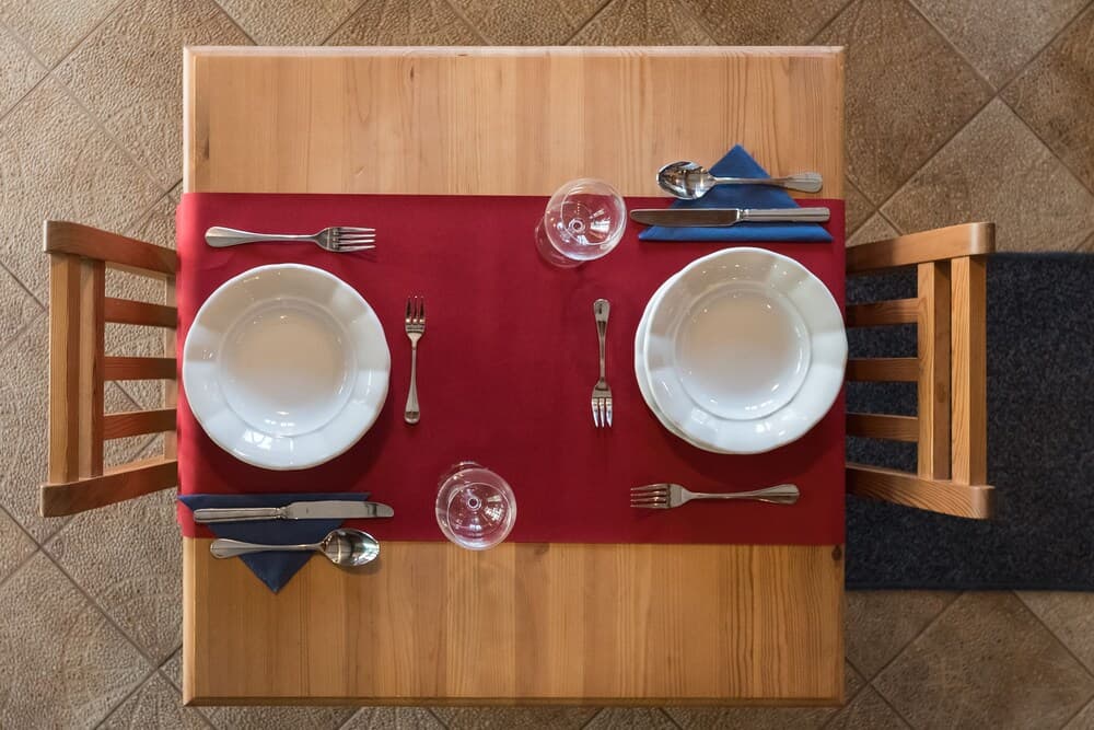 A cozy restaurant table set for two with red placemats, white plates, and neatly arranged cutlery on wooden chairs, ideal for a romantic dining experience.