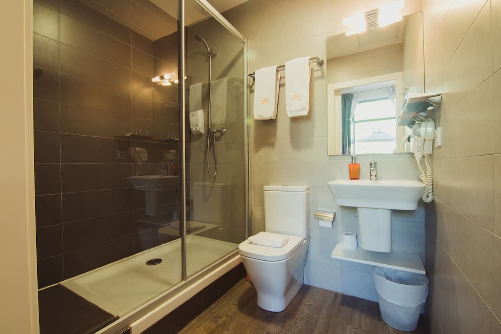 A cozy hotel bathroom features a glass-enclosed shower, white fixtures, wooden flooring, and natural light from a window, ideal for travelers seeking comfort and cleanliness.