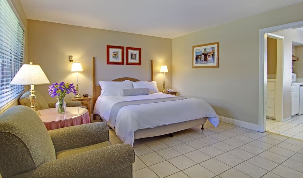 A cozy hotel room with tiled floors, a neatly made bed, soft lighting, and a small kitchenette in the background offers a comfortable stay for travelers seeking relaxation.