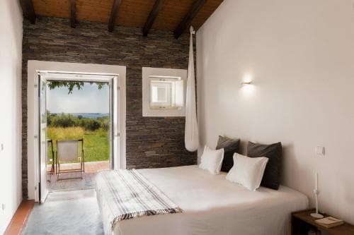 A cozy bedroom with rustic wooden beams and stone walls opens to a serene outdoor view, featuring a comfortable bed, soft lighting, and a chair on the patio, ideal for a tranquil getaway in a countryside retreat.