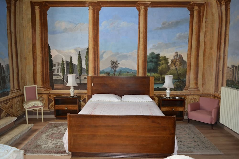 A serene bedroom with a wooden bed framed by ornate columns and a picturesque mural backdrop, featuring cozy seating and vintage decor, ideal for travelers seeking a historic and artistic retreat.