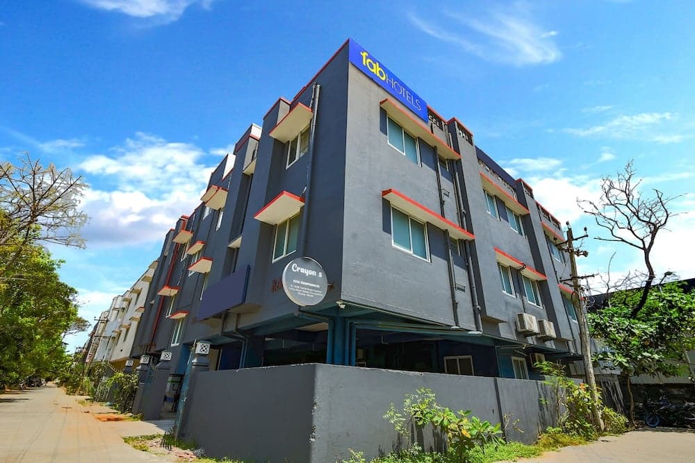A modern multi-story hotel with vibrant blue and red accents stands under a clear blue sky, surrounded by lush greenery, ideal for travelers seeking comfortable accommodations in an urban setting.
