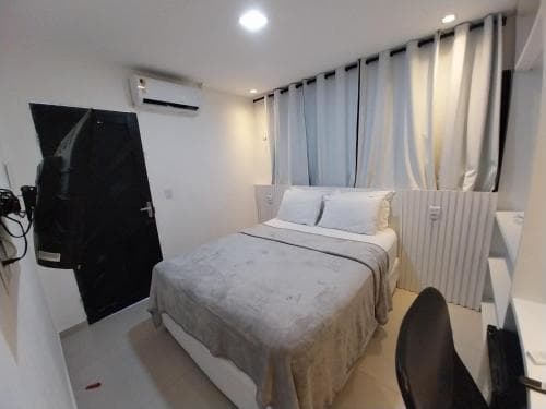 A cozy hotel room features a neatly made bed with white linens, soft curtains covering the window, and modern amenities like an air conditioner and a desk chair, ideal for travelers seeking comfort and convenience.