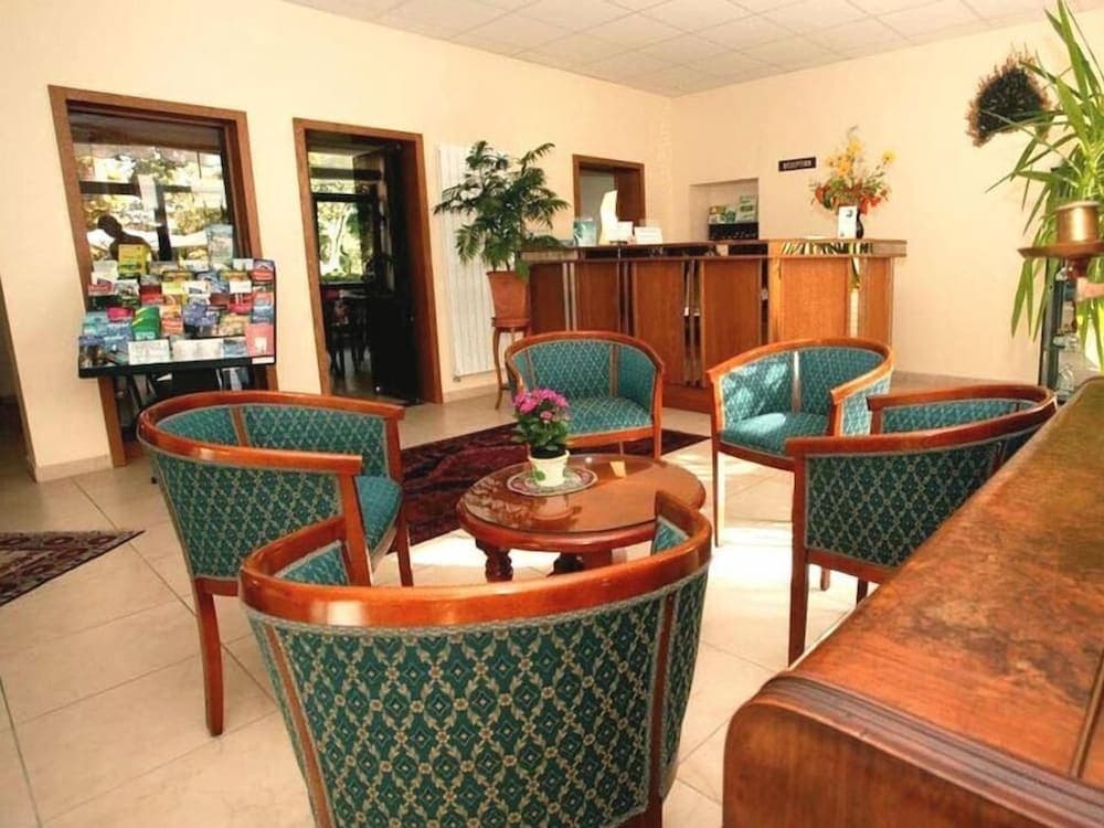 A cozy hotel lobby features patterned green chairs around a wooden table with a floral centerpiece, set against a backdrop of potted plants and a reception desk, ideal for travelers seeking a welcoming atmosphere for relaxation or business meetings.