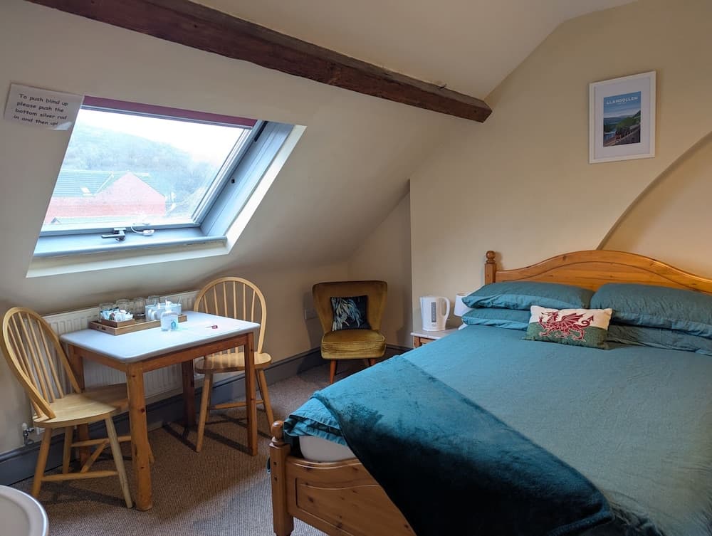 A cozy attic bedroom with a wooden bed, teal bedding, a small dining table by the window, and a scenic view of rooftops and hills, ideal for travelers seeking a charming and intimate retreat.