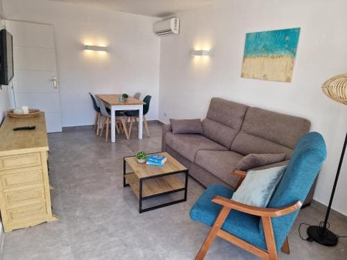 A cozy modern apartment living room features a comfortable sofa, wooden dining table, and vibrant blue armchair, creating an inviting atmosphere ideal for travelers seeking a stylish yet homey accommodation.