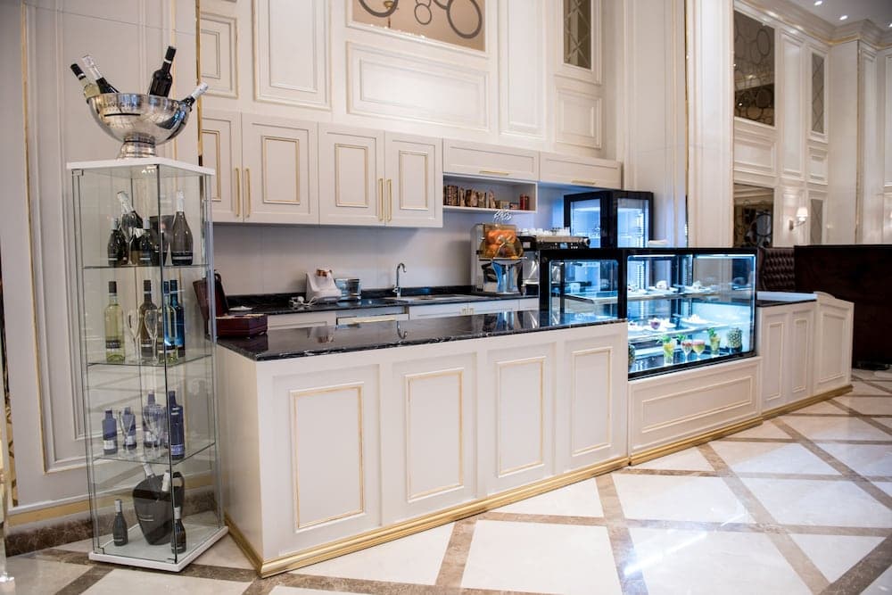 An elegant hotel bar area features a sleek white counter with gold accents, glass display cases filled with premium spirits, and a well-lit pastry display, creating an inviting atmosphere for travelers seeking a luxurious refreshment spot.