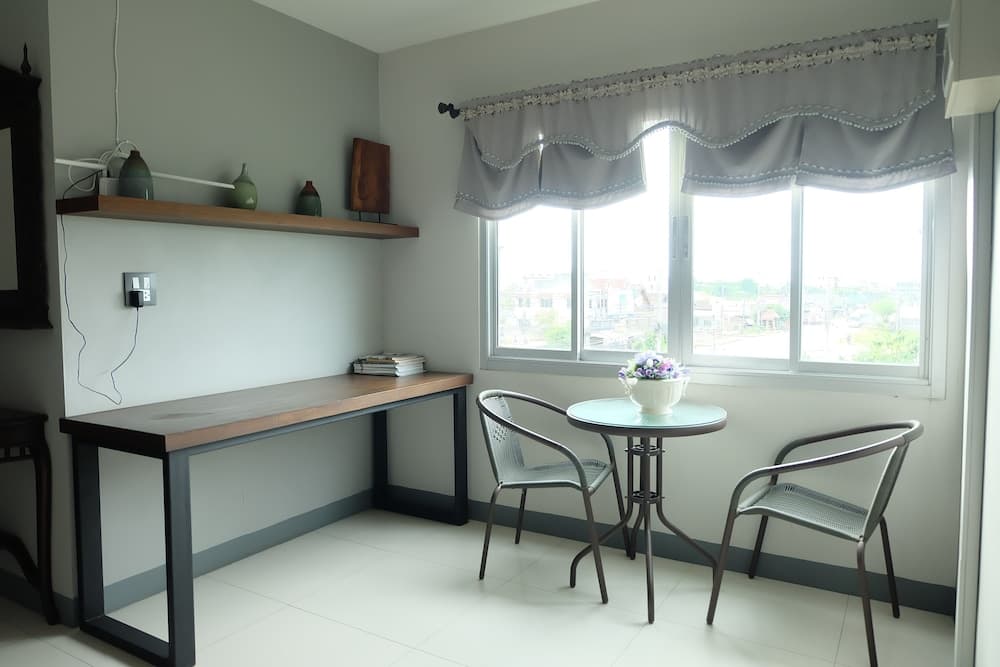 A cozy corner in a minimalist room features a wooden desk with shelves displaying vases, a small round table with chairs adorned by a flower pot, and a window with lace curtains offering a serene view, ideal for travelers seeking a peaceful workspace or relaxation spot.