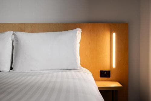 A cozy hotel room features a neatly made bed with white linens against a warm wooden headboard, illuminated by a modern vertical light, ideal for travelers seeking comfort and style.