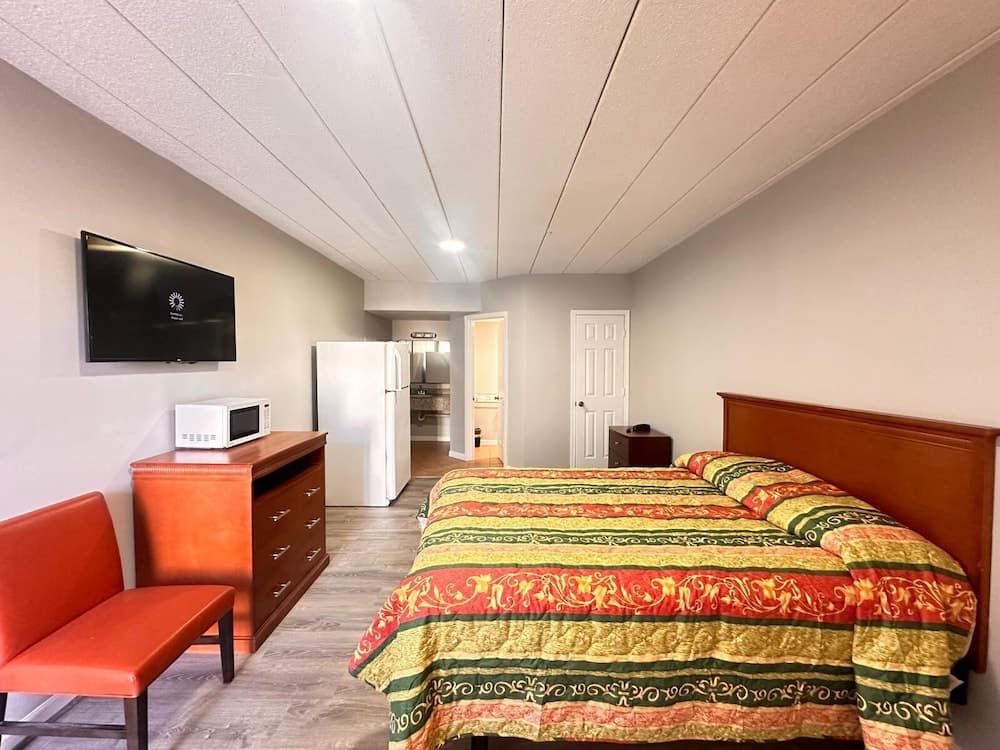 A cozy motel room features a vibrant bedspread, a mounted TV, a microwave on a wooden dresser, and a glimpse into a compact kitchen area, ideal for travelers seeking comfort and convenience.