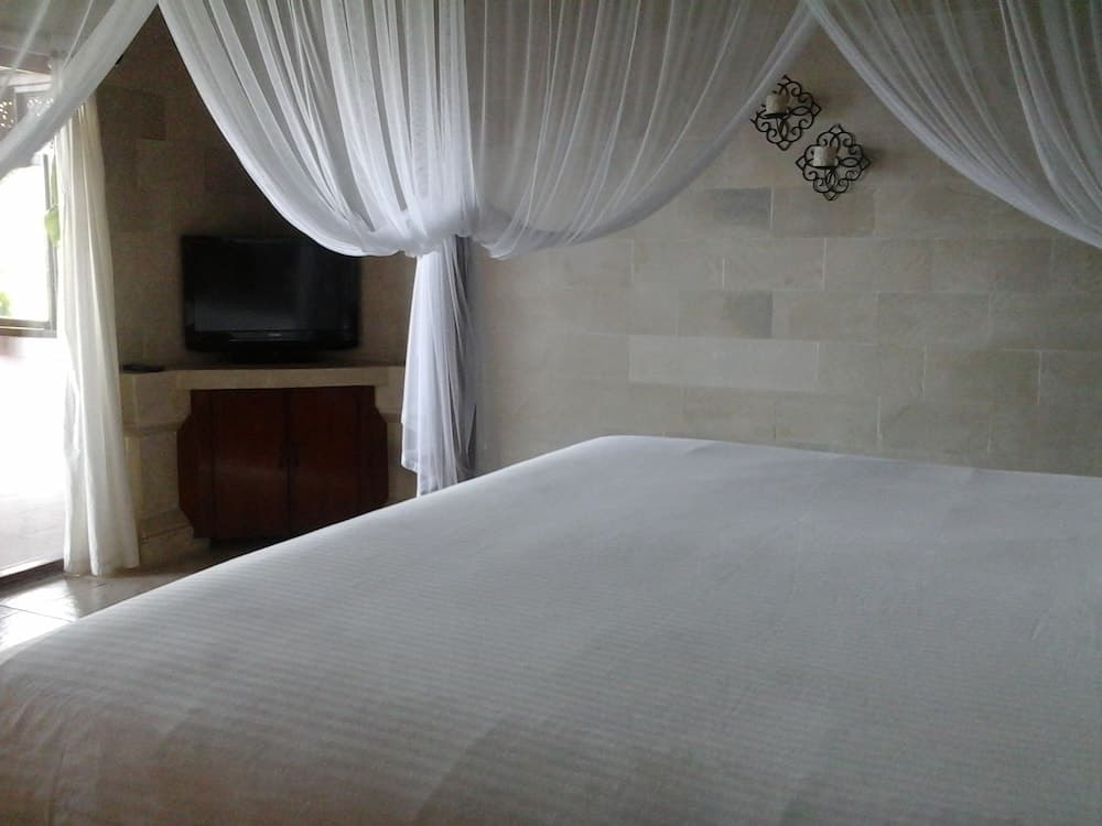 A serene hotel bedroom features a cozy bed draped in flowing white curtains, stone walls, a mounted TV, and ornate wall sconces, ideal for travelers seeking relaxation and comfort.