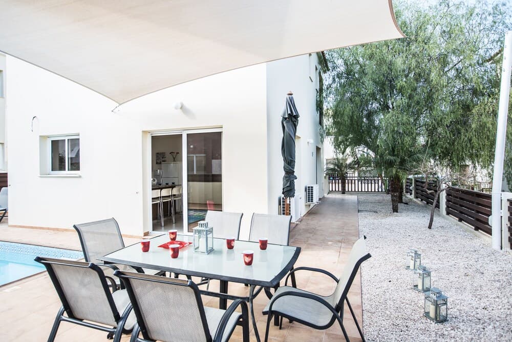 A serene private patio features a glass dining table set under a canopy, surrounded by lush greenery and a poolside ambiance, ideal for a relaxing vacation retreat.