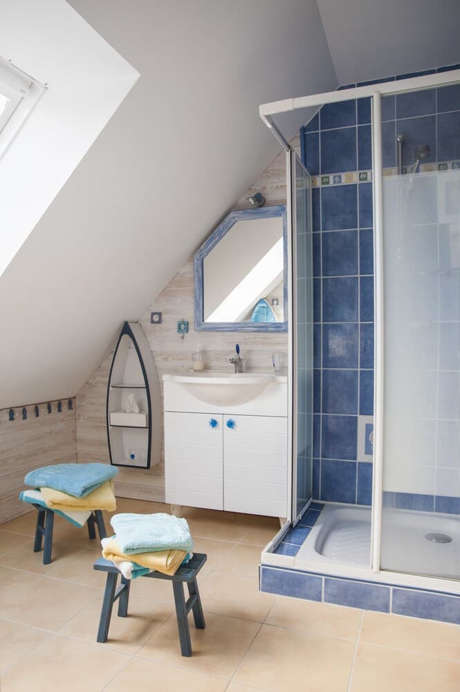 A cozy attic bathroom with blue-tiled shower, wooden accents, and neatly folded towels on stools, ideal for travelers seeking a charming and functional space.