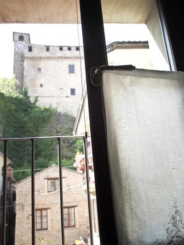 A serene view from a balcony showcases a historic stone castle perched on a hillside amidst lush greenery, framed by a partially drawn curtain, ideal for travelers seeking picturesque European charm.