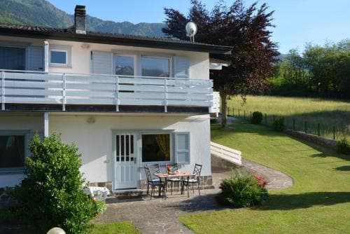 A cozy two-story house with a balcony overlooks a lush green lawn and distant mountains, creating a serene retreat ideal for a peaceful vacation getaway.