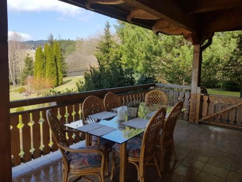 A cozy wooden balcony with a glass table and rattan chairs overlooks a serene garden, perfect for a tranquil getaway in a countryside retreat.