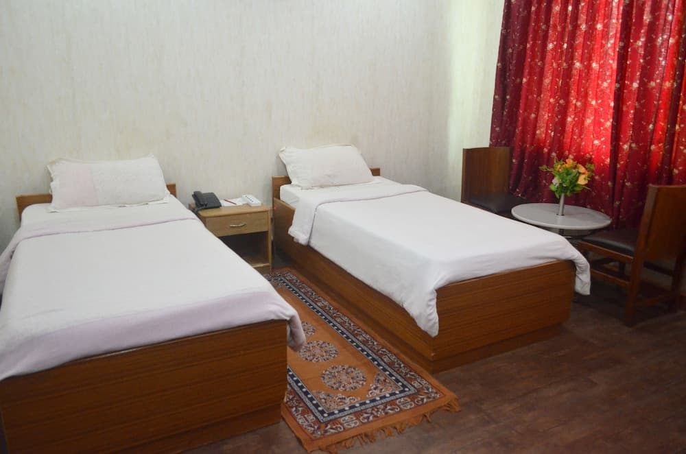 A cozy hotel room features two neatly made wooden beds with white linens, a vibrant red curtain, a small table with flowers, and a patterned rug, ideal for travelers seeking comfort and simplicity.