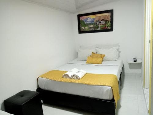 A cozy hotel room features a neatly made bed adorned with gold accents, a framed mountain landscape painting above it, and minimalistic decor, ideal for travelers seeking a simple yet comfortable stay.