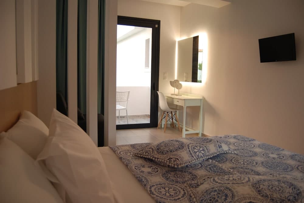 A cozy hotel room features a neatly made bed with blue patterned bedding, a bright balcony view through glass doors, a stylish vanity with a mirror, and a wall-mounted TV, ideal for travelers seeking comfort and relaxation.