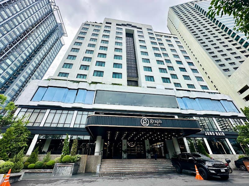 A modern urban hotel entrance with sleek glass architecture and lush greenery sets a welcoming tone in Bangkok's bustling cityscape, ideal for travelers seeking upscale accommodations.