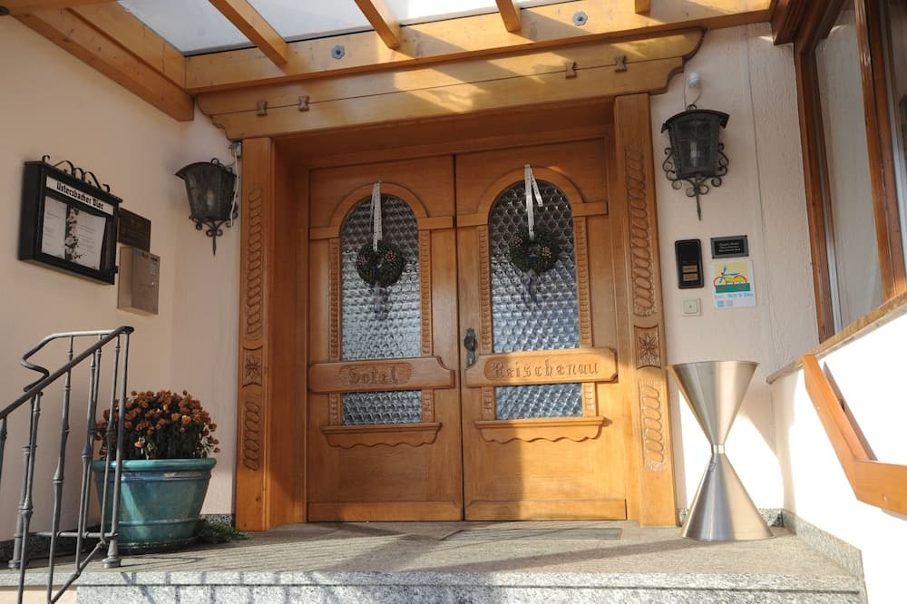 A charming hotel entrance features ornate wooden doors adorned with glass panels and wreaths, flanked by lanterns and a potted plant, inviting travelers to a cozy stay in an alpine setting.