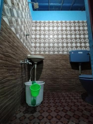A compact bathroom with patterned tiles, a blue toilet, and a green bucket under a sink, ideal for travelers seeking basic facilities in budget accommodations.