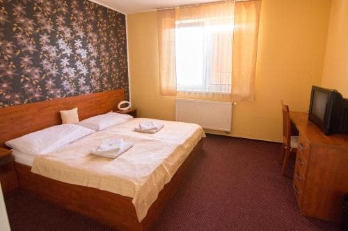 A cozy hotel room features a neatly made bed beside a window with sheer curtains, a small desk with a CRT TV, and a patterned wall, ideal for budget travelers seeking comfort.