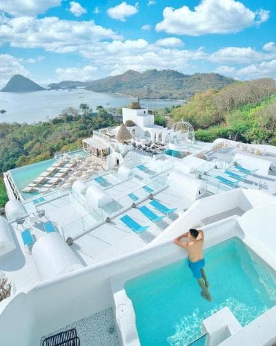 A serene rooftop pool area with lounge chairs overlooks lush green hills and a tranquil sea under a bright blue sky, ideal for luxury resort relaxation and scenic photography.