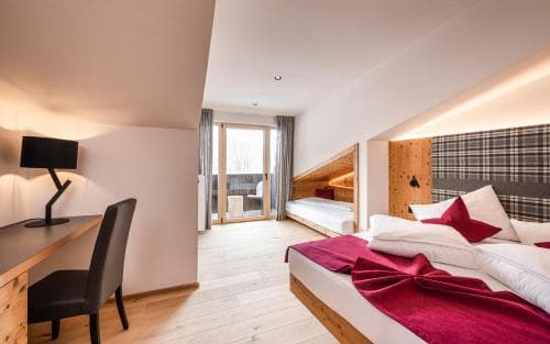 A cozy hotel room with wooden accents, a neatly made bed adorned with red pillows, a desk with a modern lamp, and a balcony offering a serene retreat, ideal for travelers seeking comfort and relaxation.
